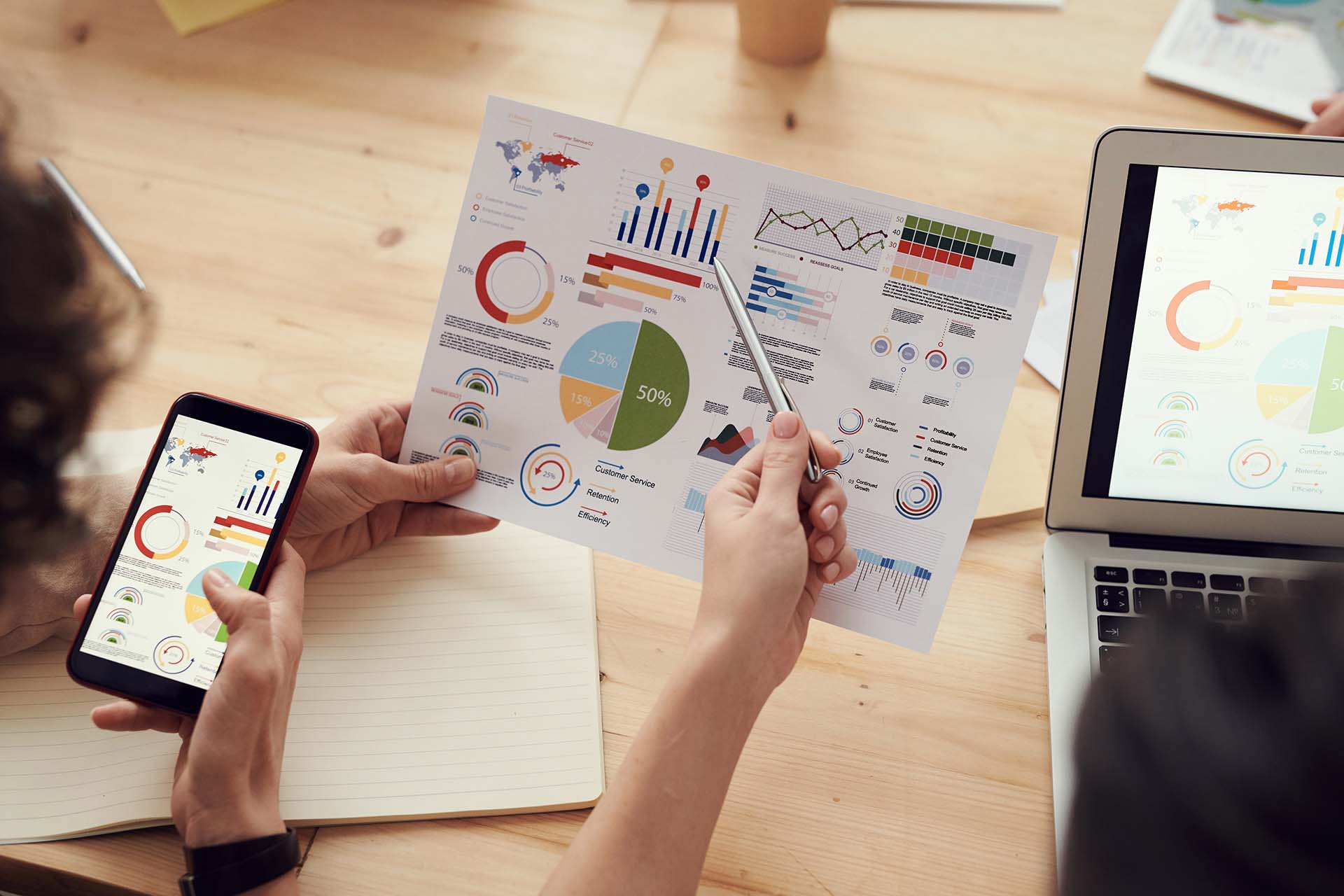 Two people review colorful business charts and graphs on paper and a smartphone at a wooden table. A laptop and notepad are also visible. One person points at the paper with a pen. The setting appears to be a collaborative work environment.