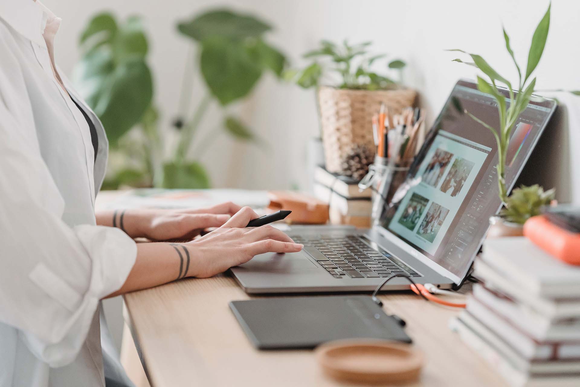Person working on a laptop at a desk, using a stylus. The workspace is decorated with potted plants, books, and stationery. A laptop screen displays various images, and a graphic tablet lies nearby.