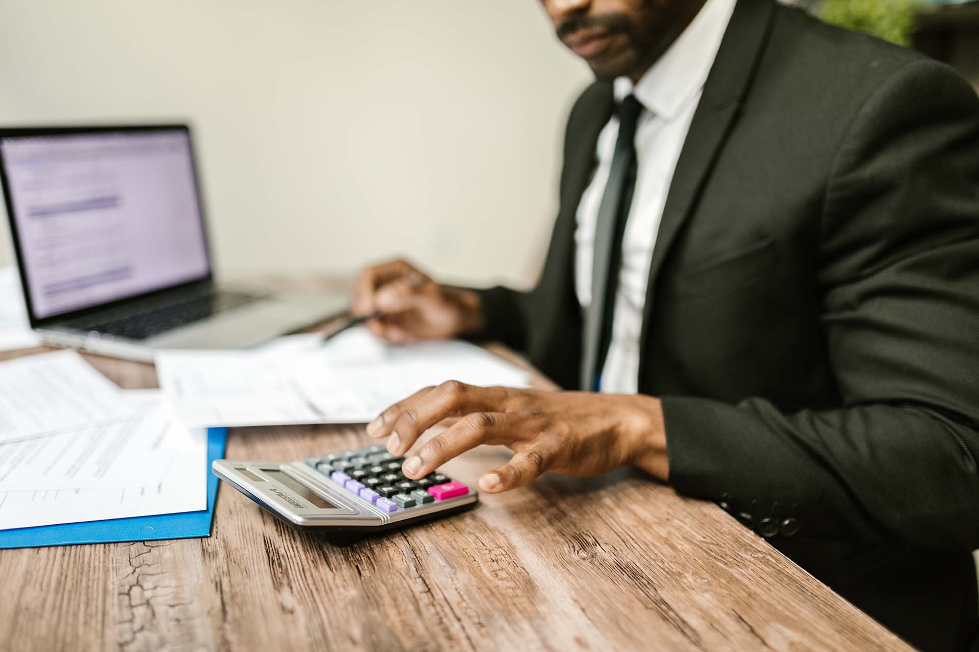 A person in a suit uses a calculator at a wooden desk with documents and a laptop. The individual is focused on paperwork, suggesting a business or financial setting. The laptop screen is partially visible.